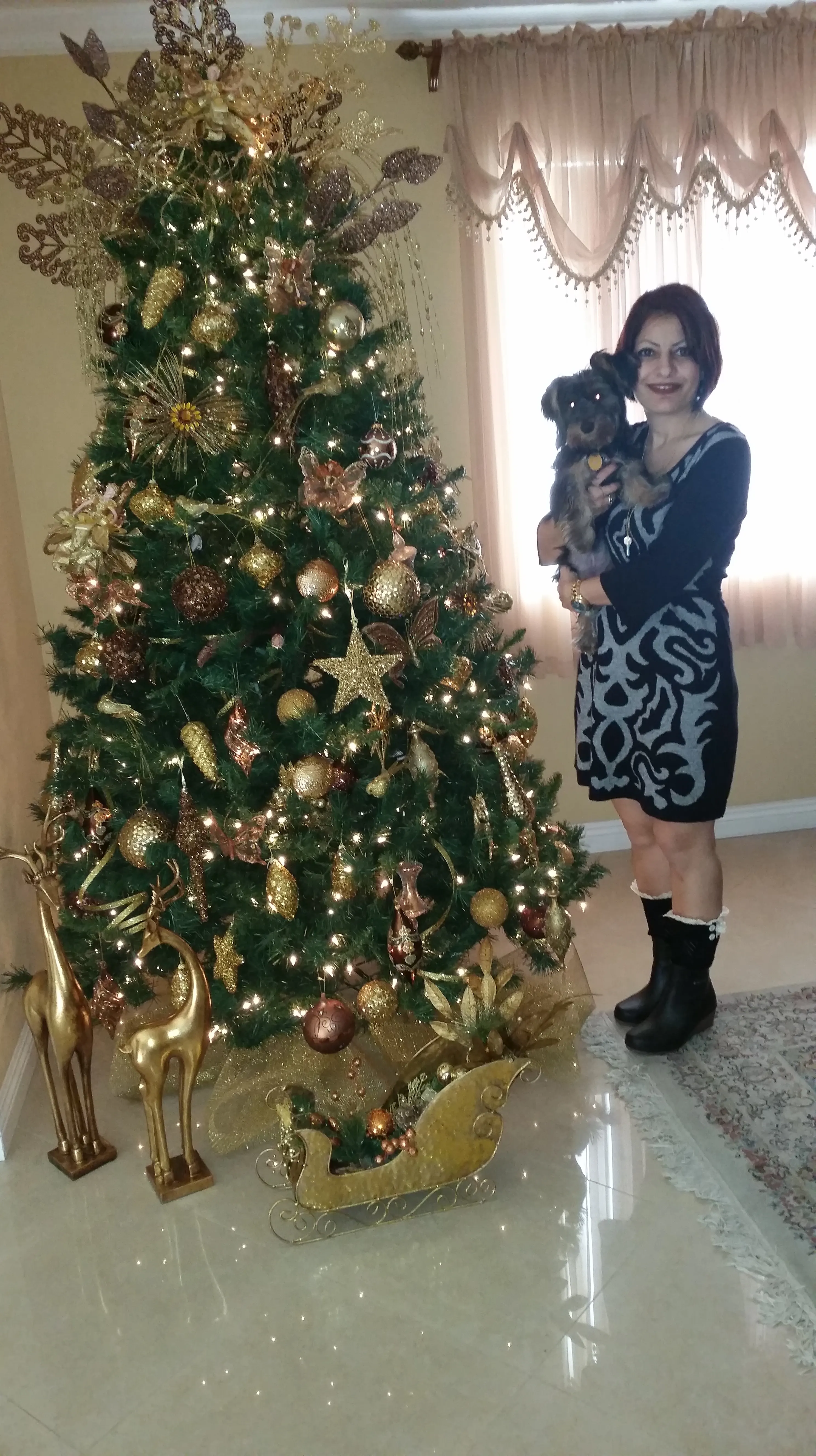A woman in a patterned dress holding Pearl as a puppy beside a tall Christmas tree decorated in gold.
