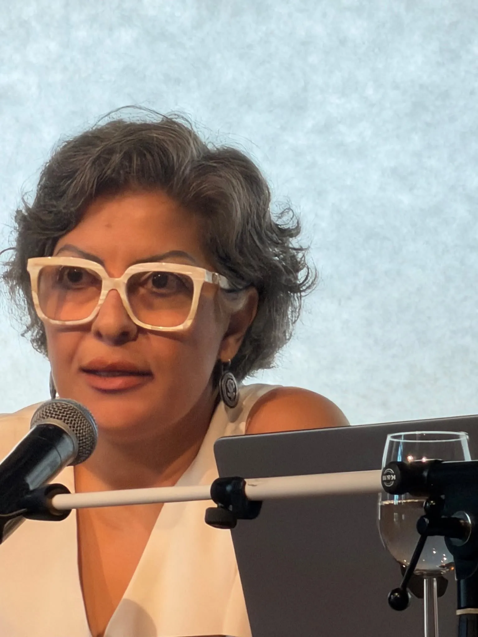 Claudia Yaghoobi seated at a table, speaking into a microphone. She has short curly gray hair and wears large white-framed glasses with a white sleeveless top. An open laptop stands beside her.