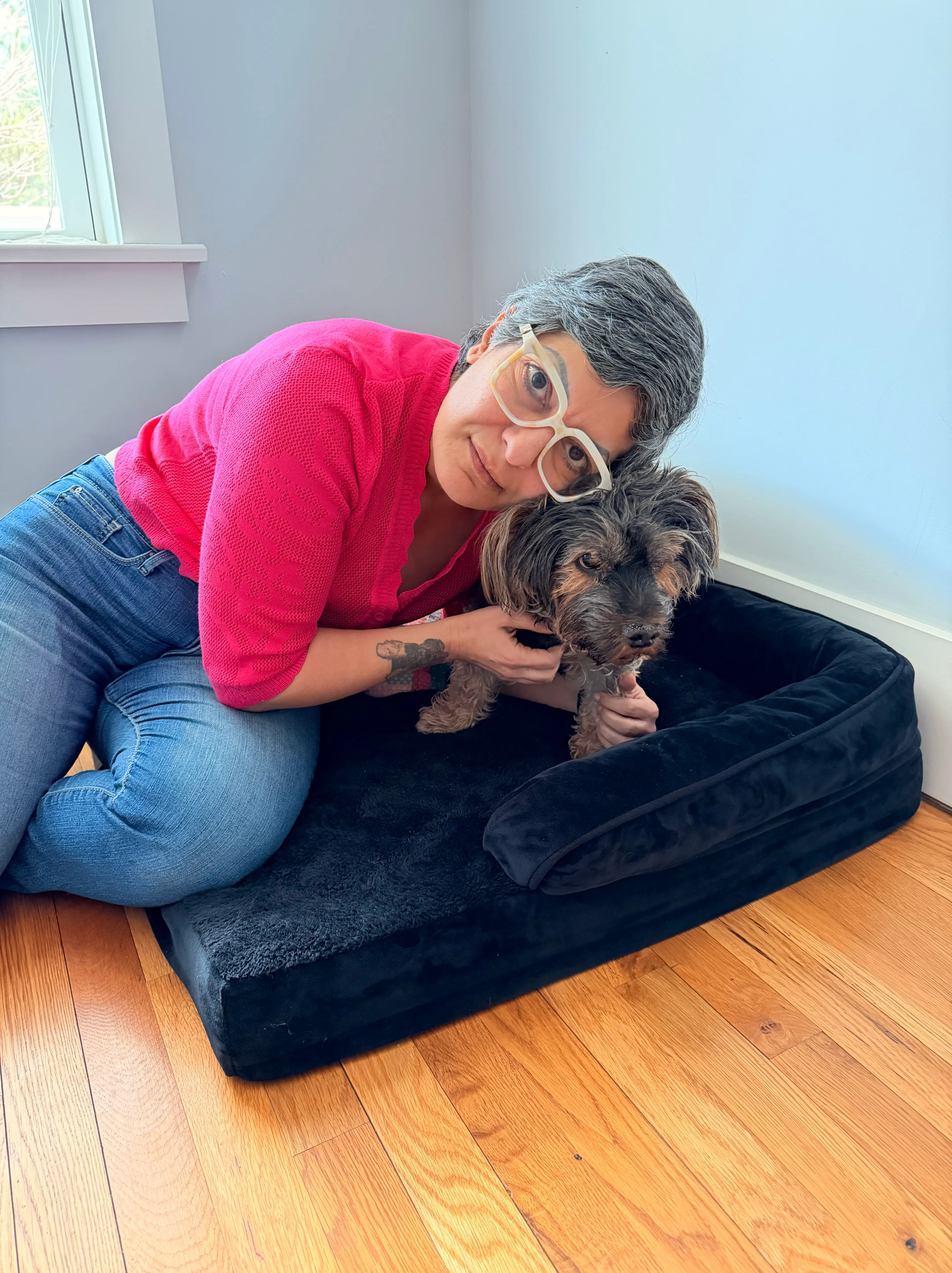 Claudia kneeling beside a navy dog bed in the corner of a room, her face close to Pearl, who sits on the bed.