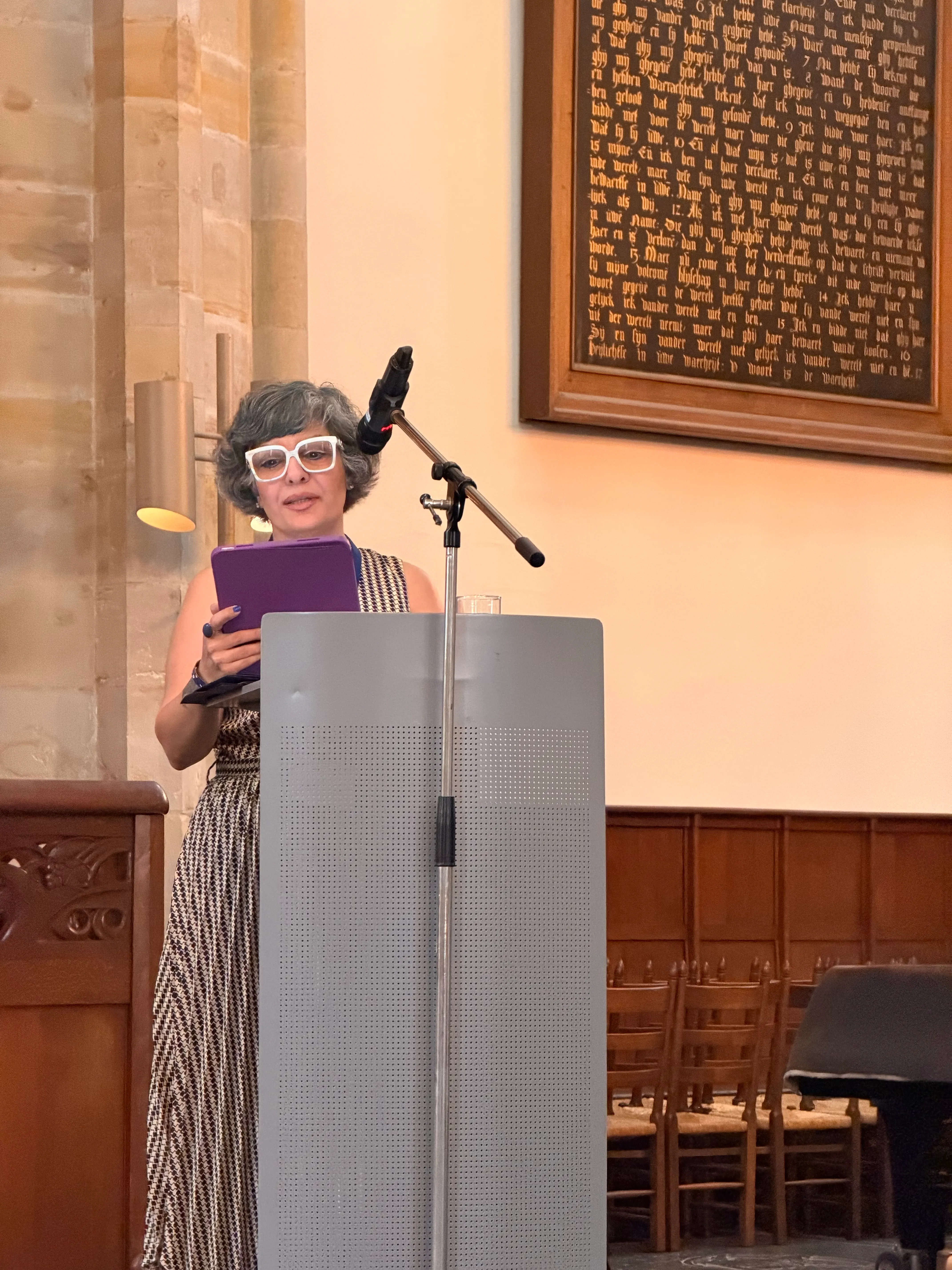 Claudia Yaghoobi speaking at a lectern with a microphone, holding a purple tablet. Behind her are wood paneling, a stone pillar and a large framed inscription.