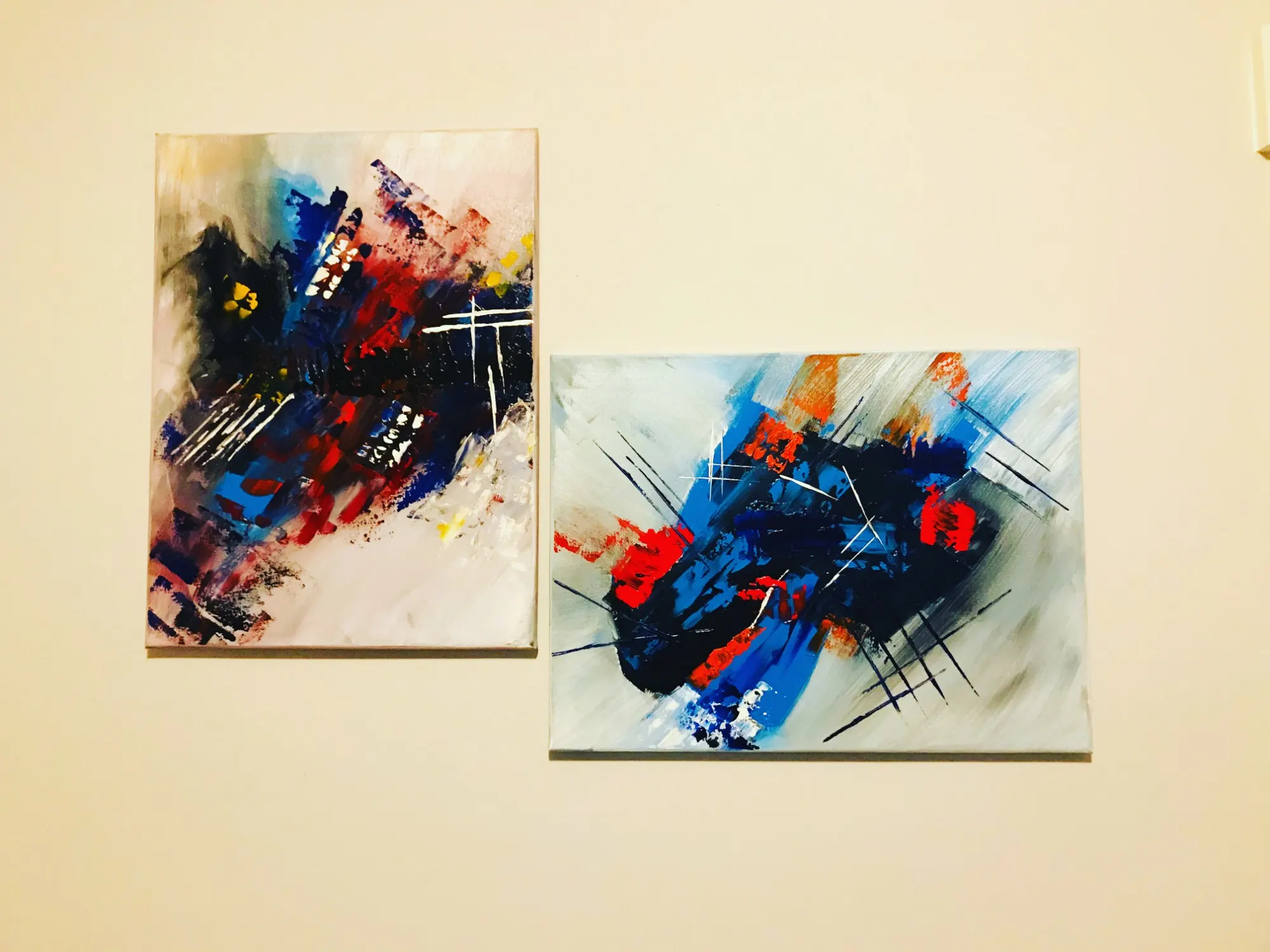 Two abstract paintings hung side by side on a cream wall. The taller canvas on the left layers navy, red and black strokes over a pale ground with flecks of yellow. The wider canvas on the right sets a dark navy mass with red patches against pale gray, crossed by thin diagonal lines.