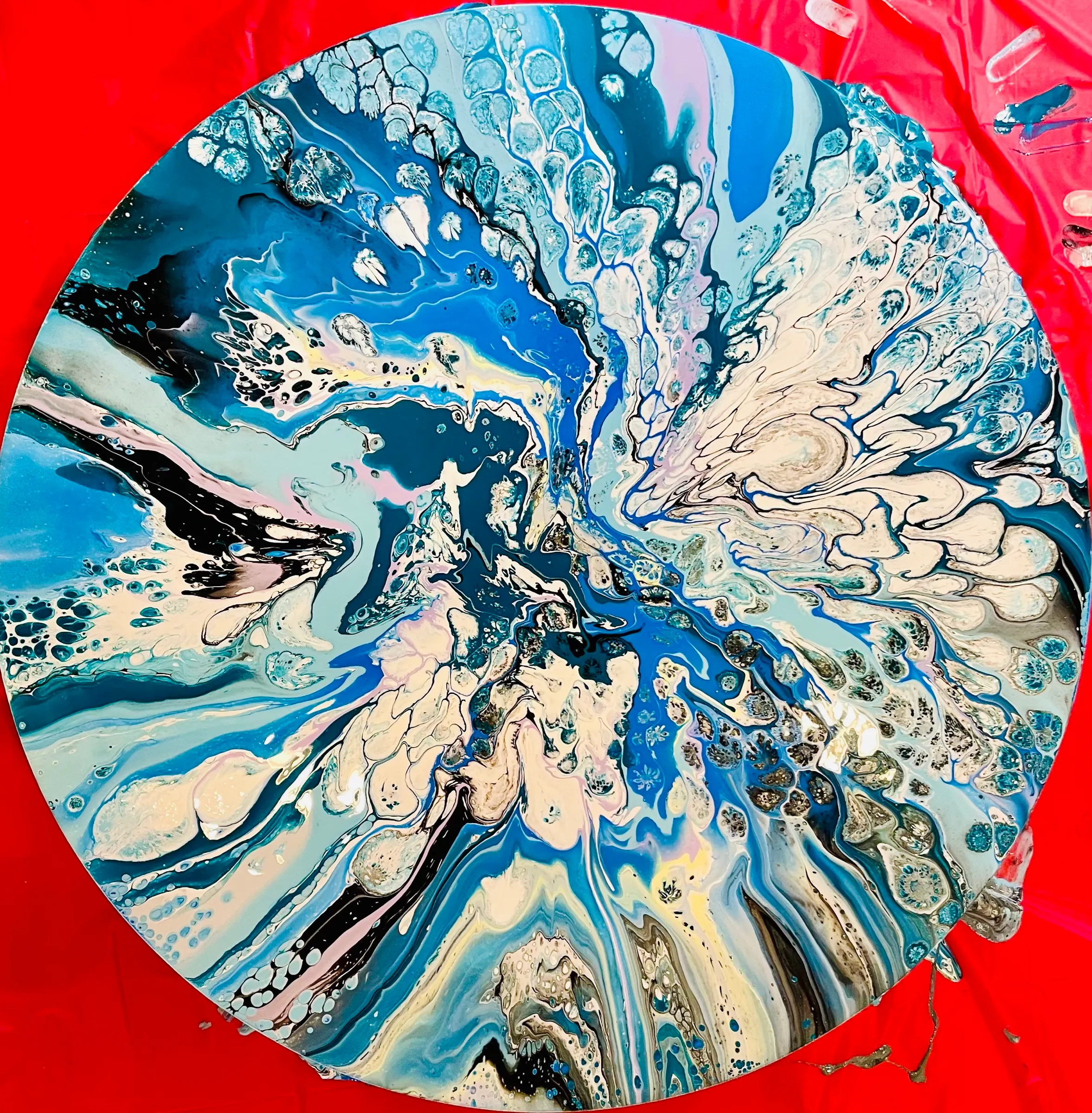 A round abstract poured painting of blue, black and white swirls with cream cells, photographed on a red cloth.